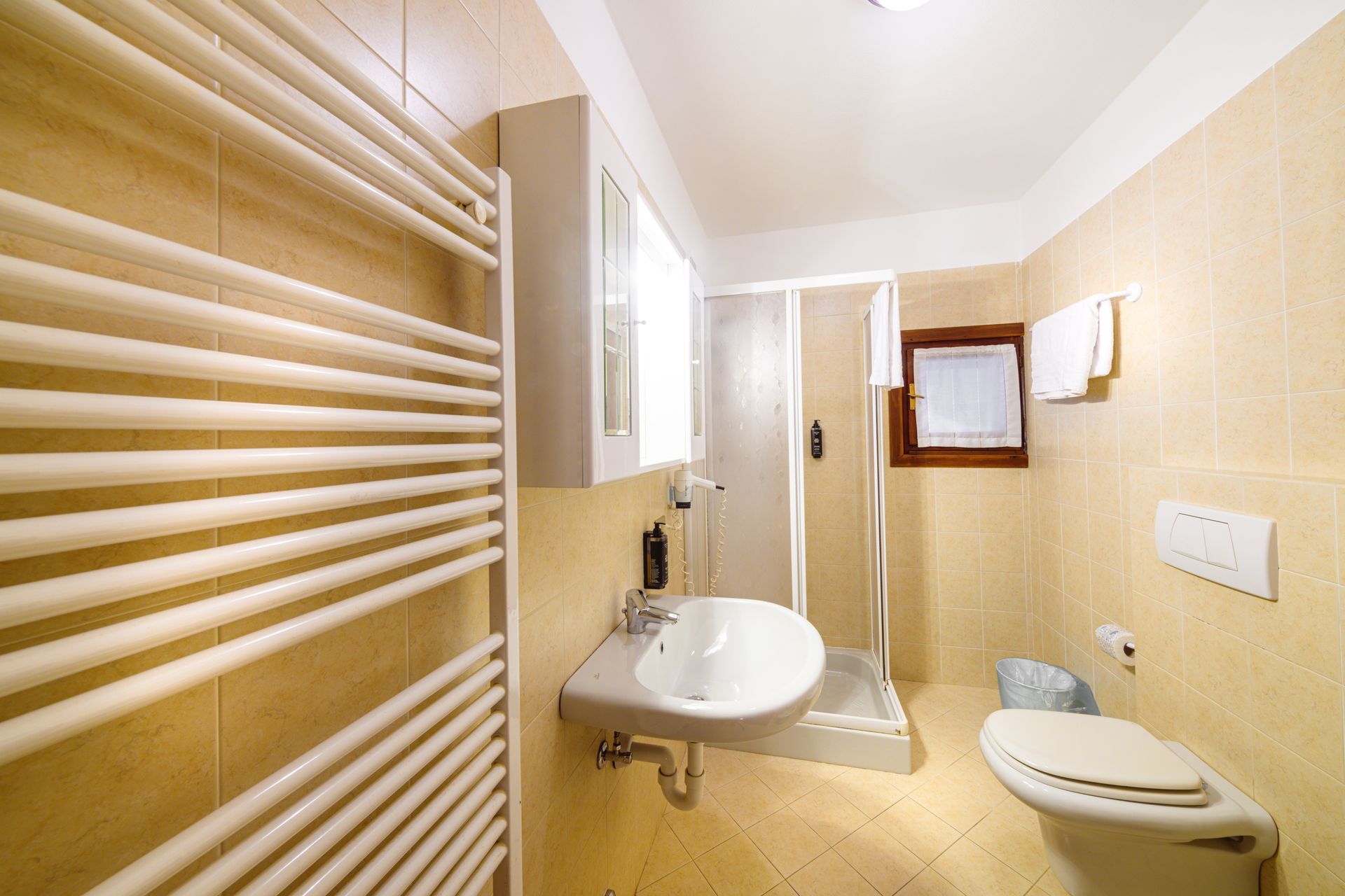 Piccolo bagno beige con lavandino, doccia, WC, termoarredo e finestra.