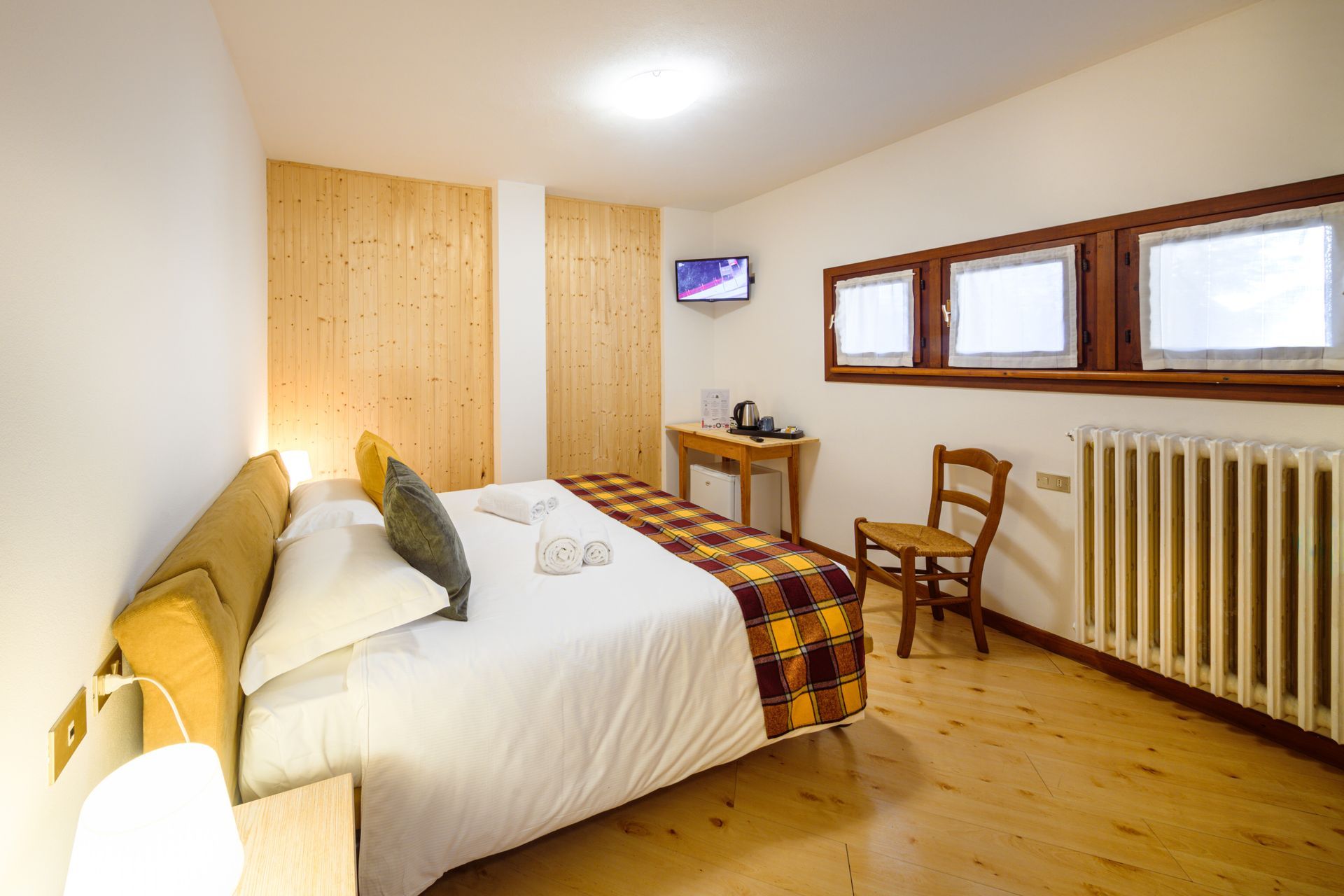 Accogliente camera da letto con letto, mobili in legno, sedia da scrivania, TV a parete e tre finestre.