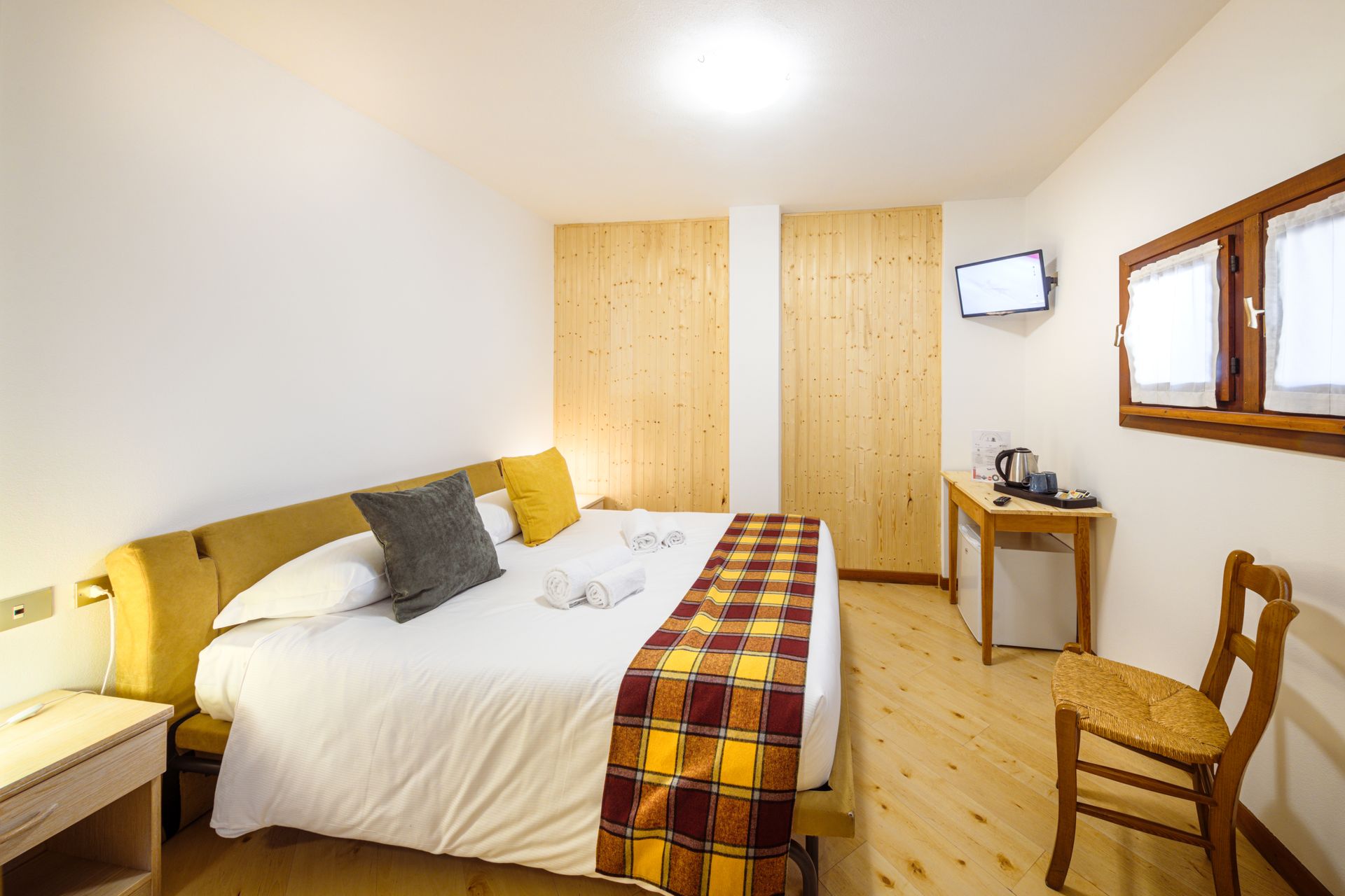 Camera da letto luminosa con letto bianco, dettagli gialli, mobili in legno e una finestra sulla destra.