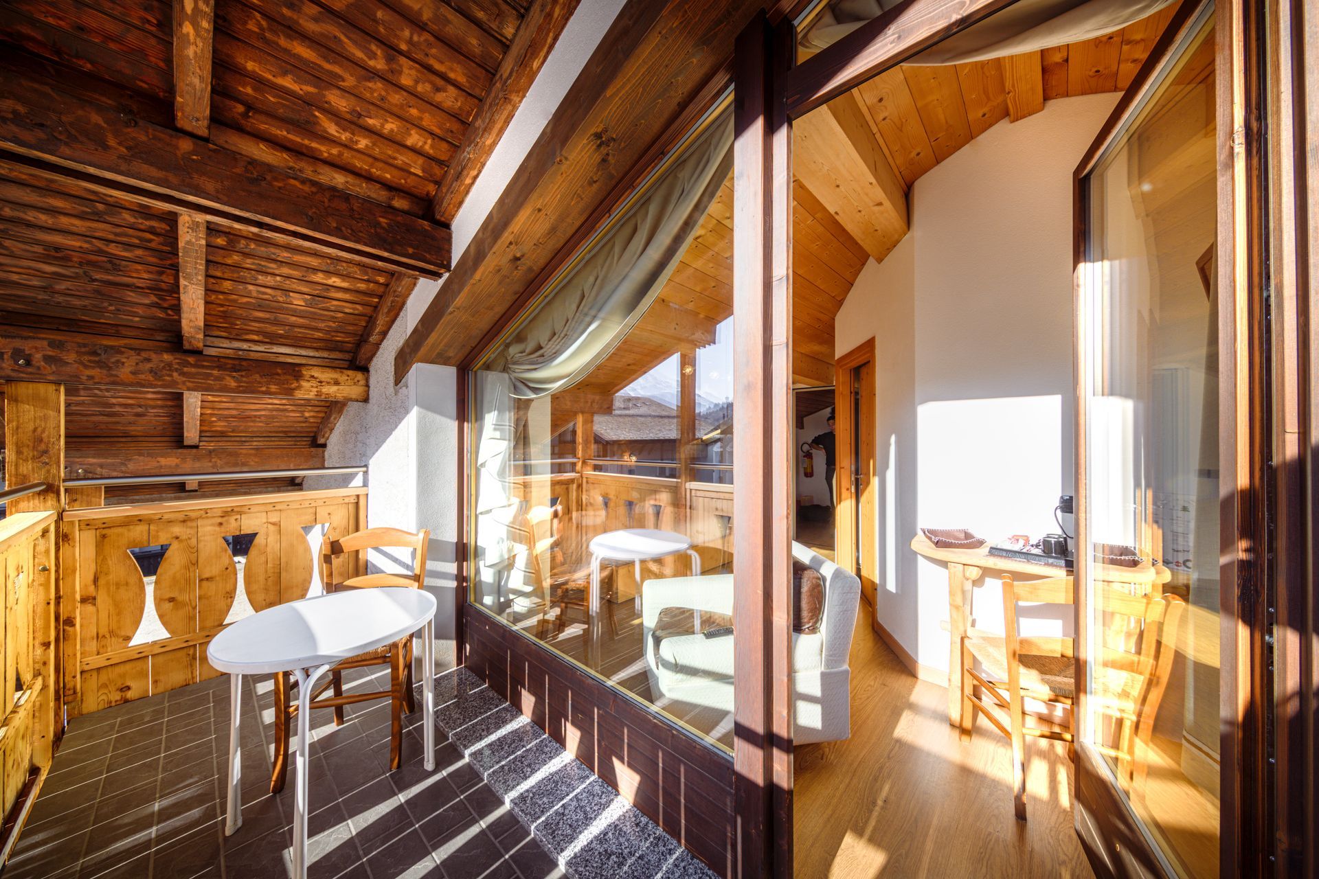 Balcone in legno con tavolo e sedie accanto a porte finestre, con vista panoramica sulle montagne.