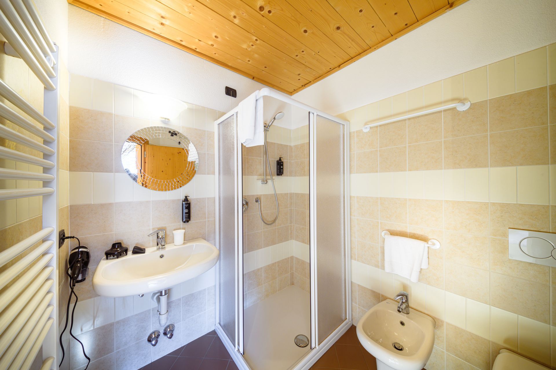 Bagno luminoso con box doccia, lavabo, specchio, WC e pareti rivestite con piastrelle beige sotto un soffitto in legno.