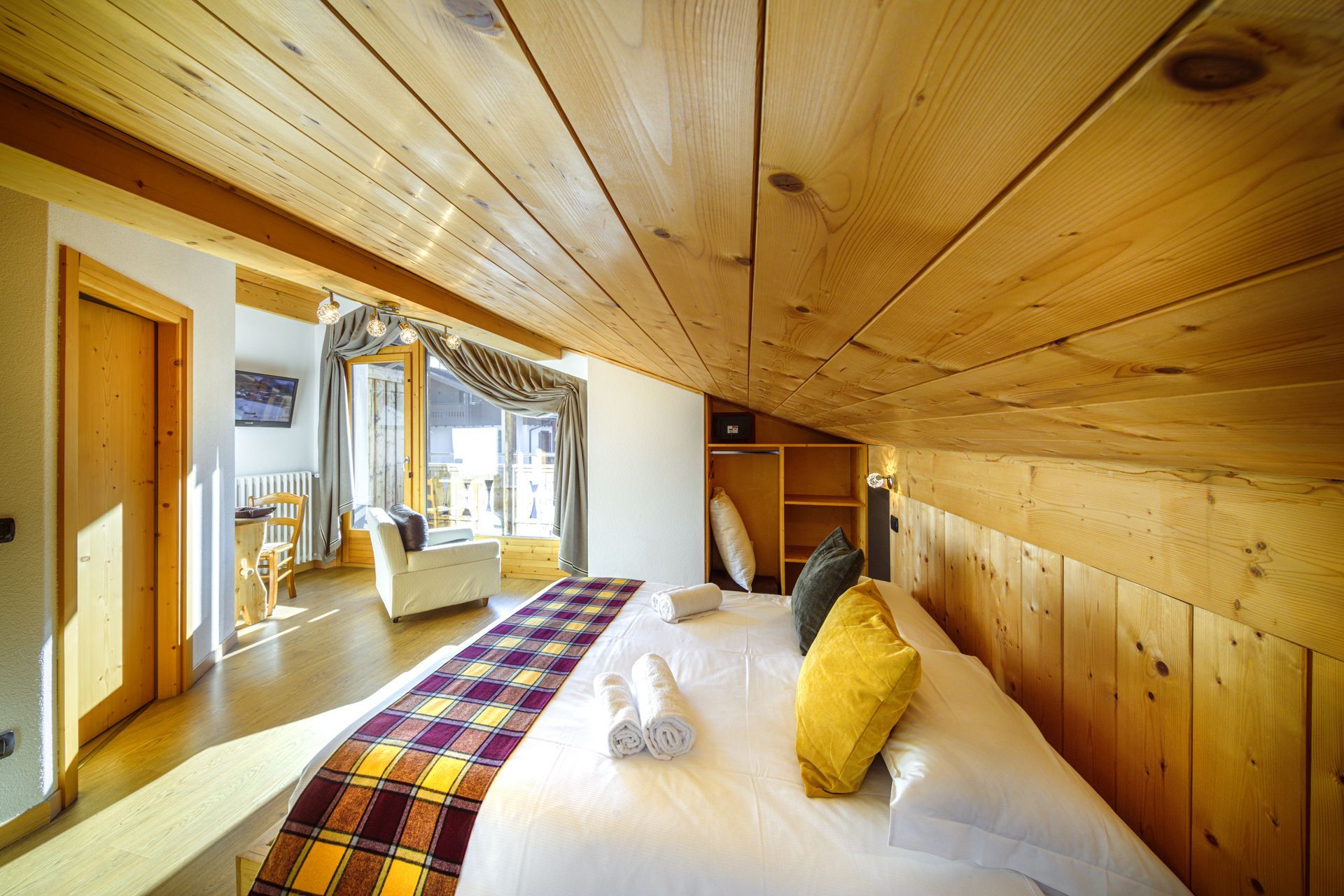 Luminosa camera da letto mansardata con pareti in legno, soffitto spiovente e un letto con coperta a quadri e cuscino giallo.