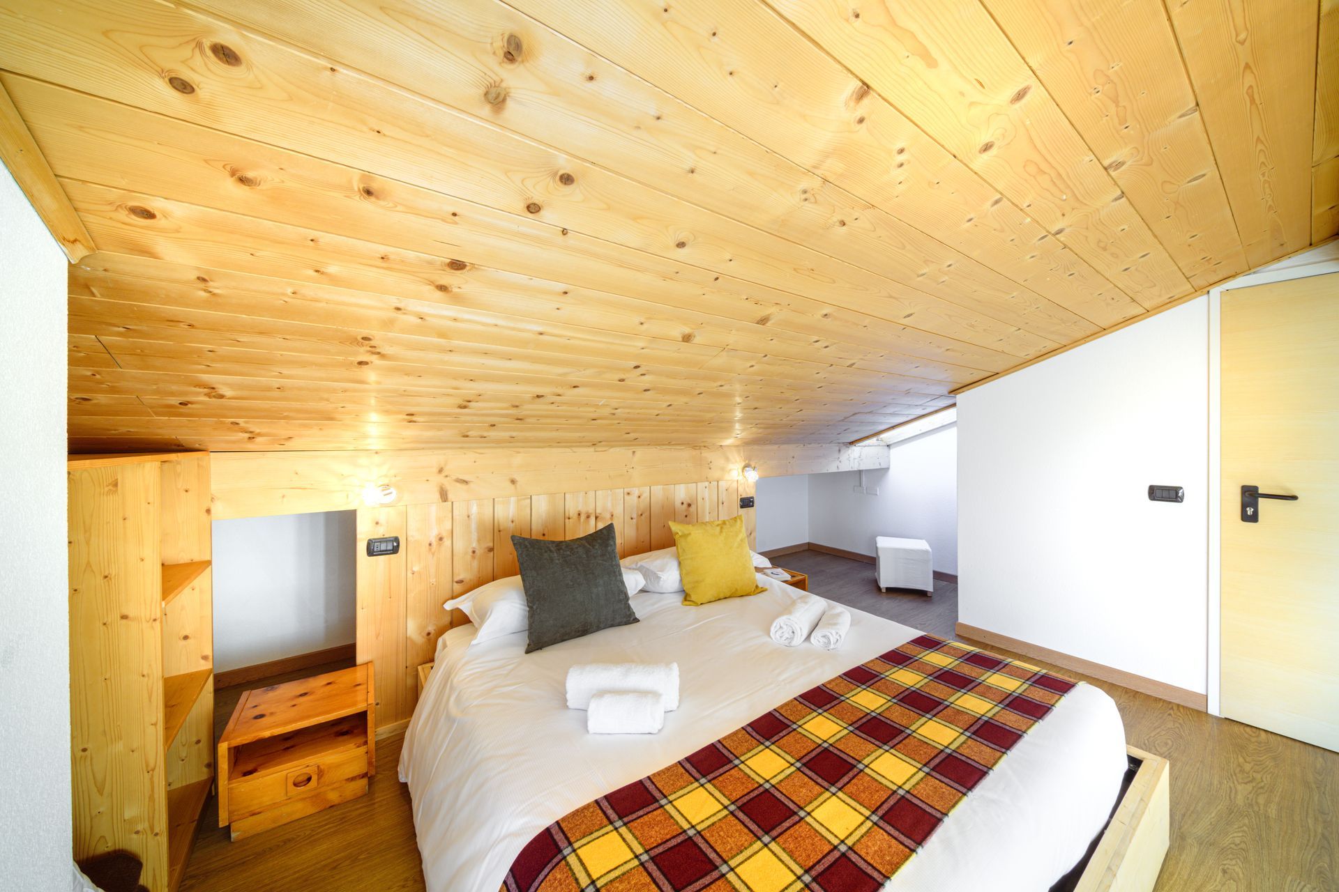 Accogliente camera da letto mansardata in legno con letto, coperta a quadri e lucernario.