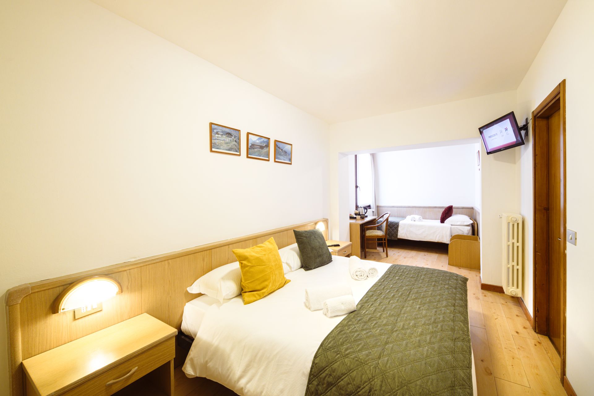 Luminosa camera d'albergo con letto, lampada da comodino, quadri alle pareti, scrivania e porta aperta verso un'altra stanza.