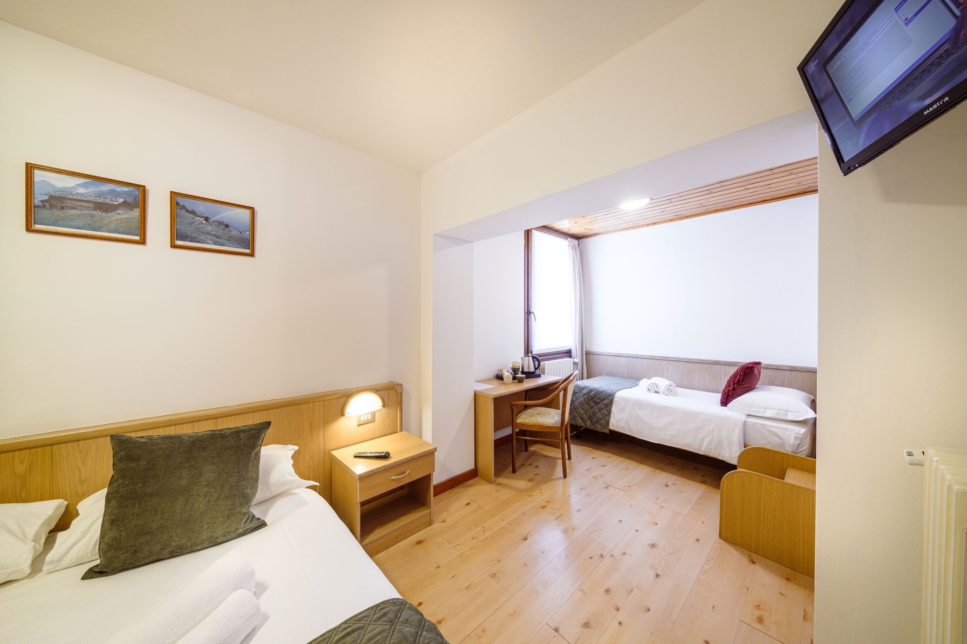 Luminosa camera d'albergo con due letti singoli, pavimento in legno, scrivania vicino alla finestra e TV a parete.