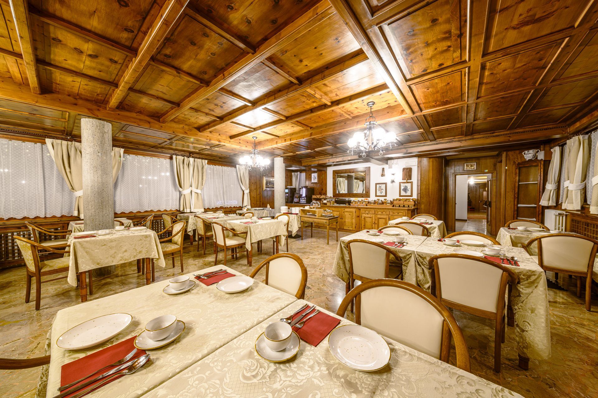 Sala da pranzo rustica vuota con soffitto in legno, tovaglie bianche e tavoli apparecchiati con piatti.