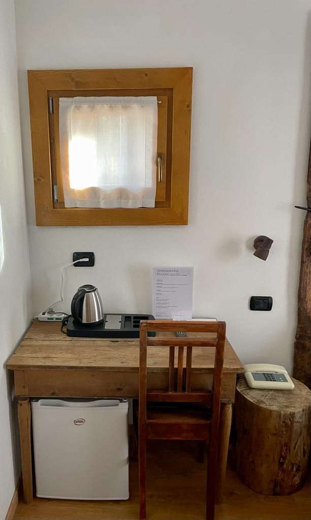 Piccola camera d'albergo con scrivania, sedia, bollitore, mini-frigo e specchio a parete.