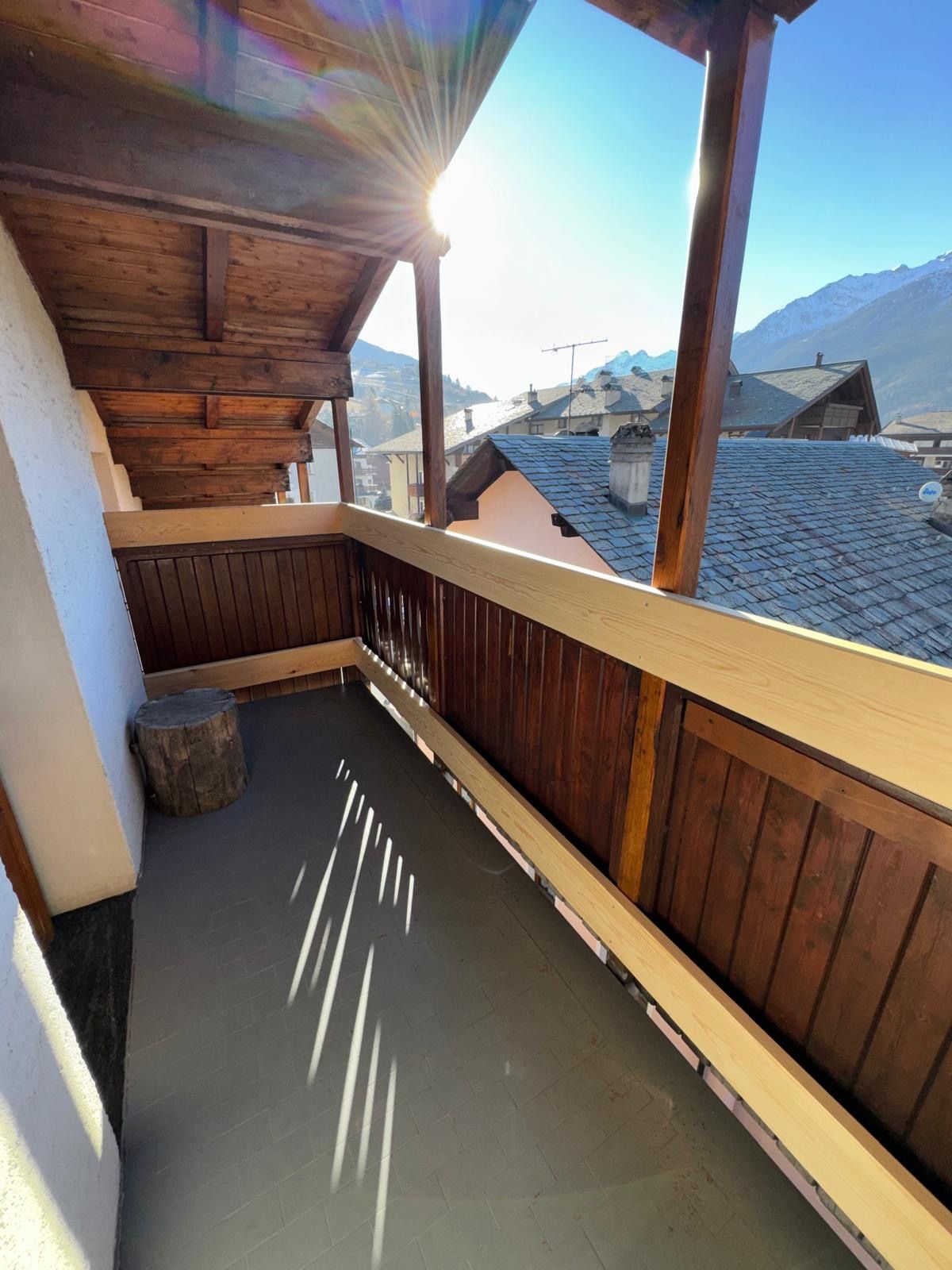 Balcone soleggiato con ringhiera in legno, piccola panchina e tetti di montagna sullo sfondo.