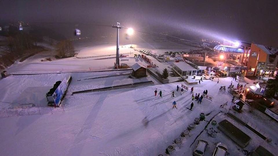 Stazione sciistica innevata al crepuscolo con piste illuminate, sciatori ed edifici che brillano sullo sfondo.