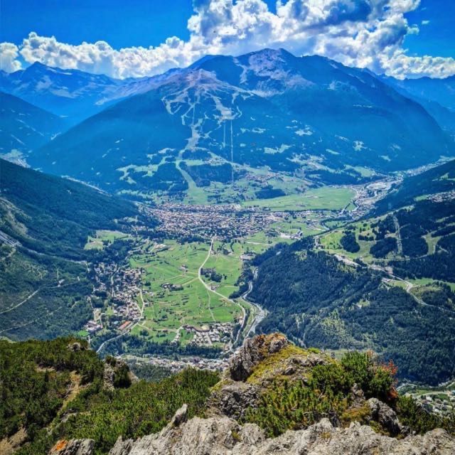 Valle di montagna con un paese, campi verdi e strade tortuose sotto cime azzurre e nuvole.