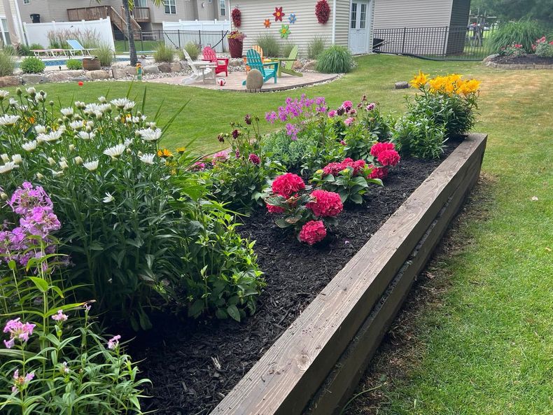 Colorful flower bed with pink, white, and yellow blooms along a raised garden border in a grassy yard