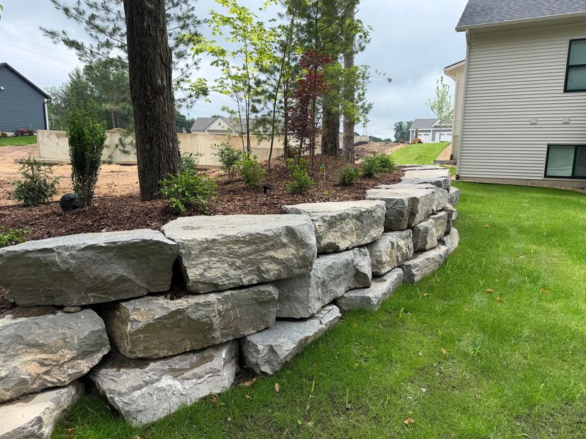Stone retaining wall edging a landscaped yard beside a house, with grass and trees.