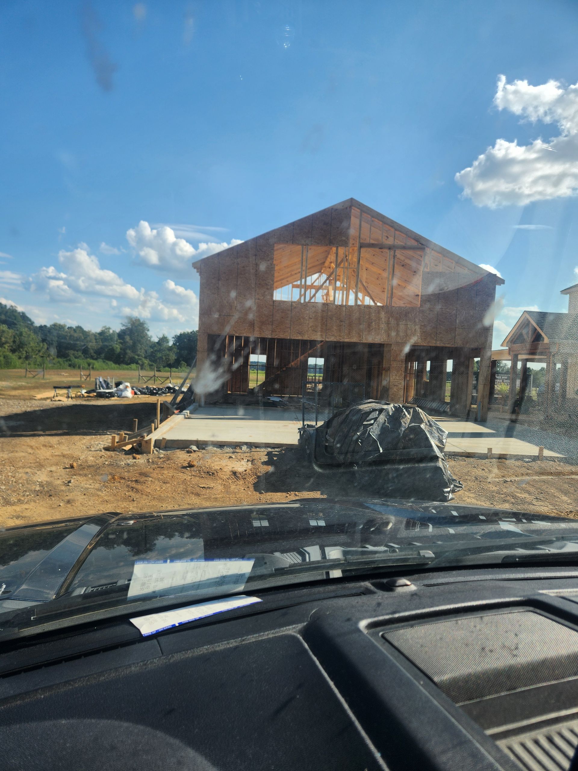 Dirt lot with a large wooden building under construction, seen from inside a car on a sunny day.