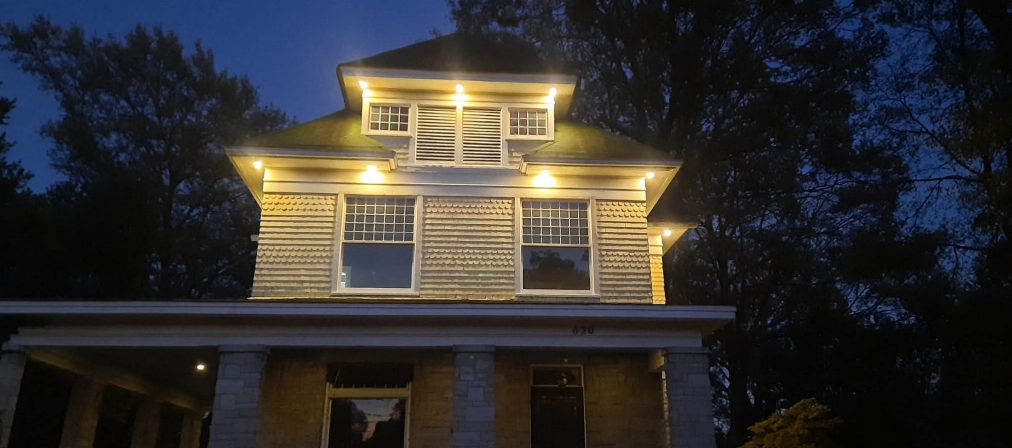 Illuminated two-story house at night with warm yellow exterior lights and dark trees behind it