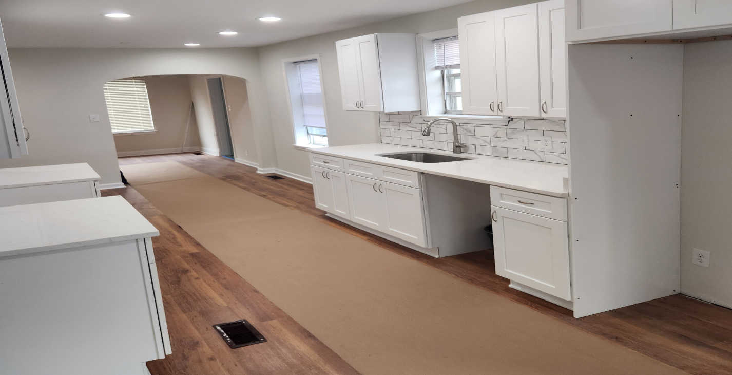 Empty renovated kitchen with white cabinets, island, and brown flooring in a long open room