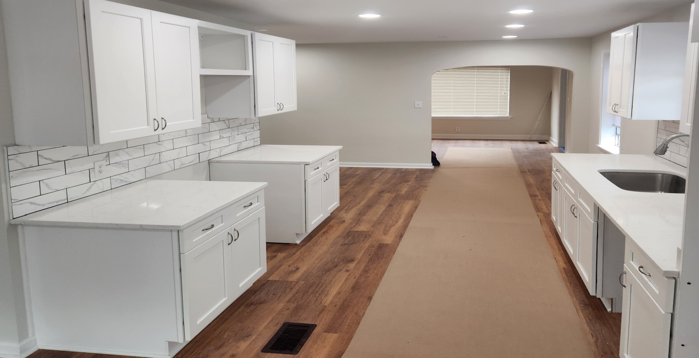 Empty kitchen under renovation with white cabinets, wood floor, and beige carpet runner