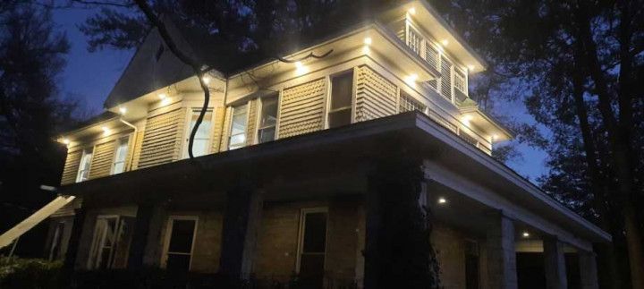 A house at dusk with warm exterior lights illuminating the upper porch and eaves.