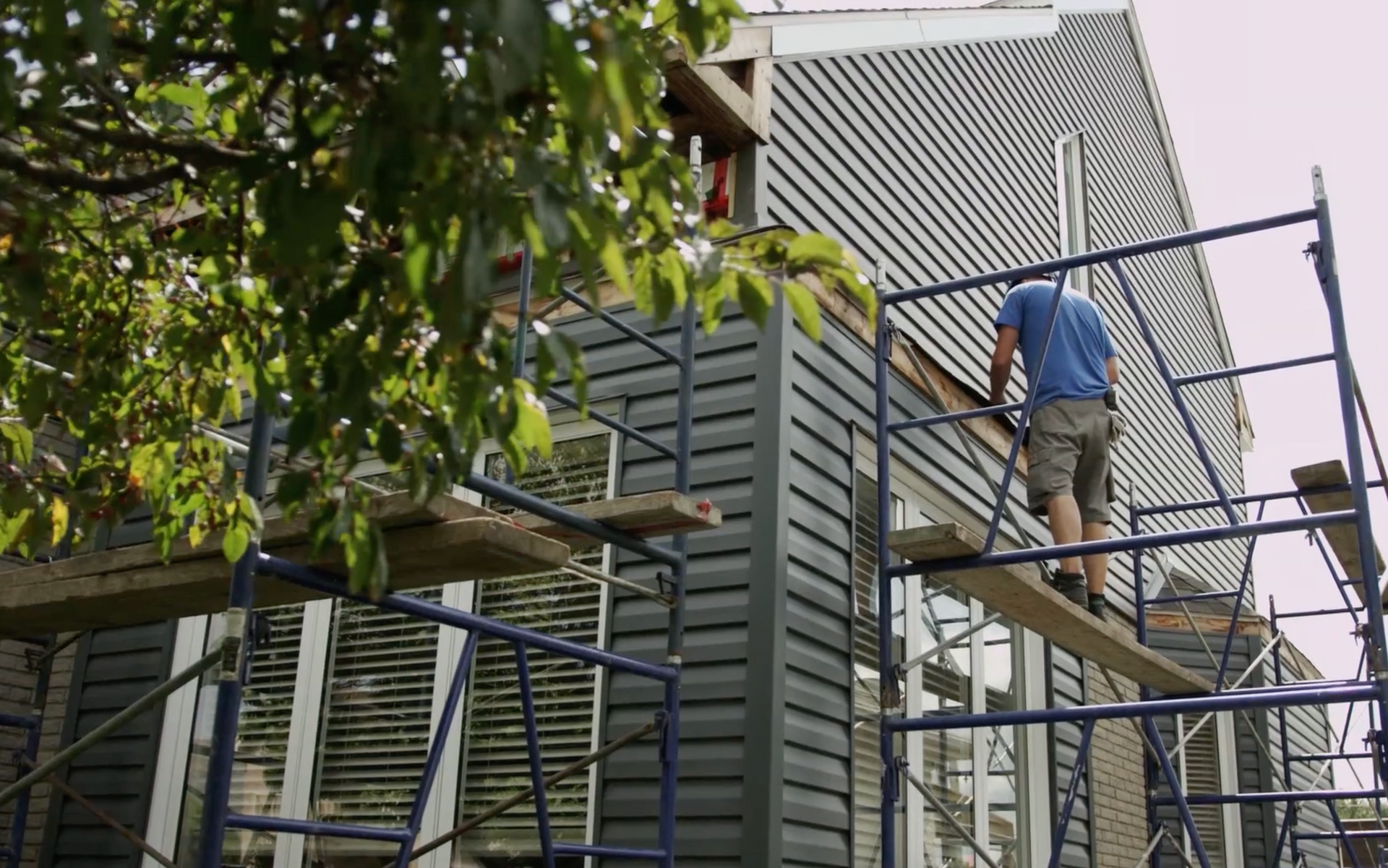 Ouvrier du bâtiment sur un échafaudage, installant un revêtement foncé sur une maison.