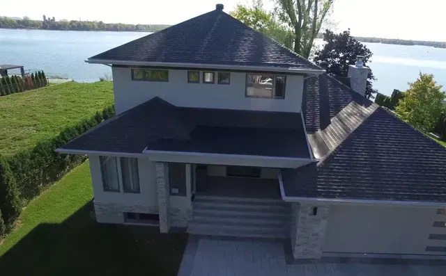 Maison à deux étages avec toit gris, face à un lac. Garage, fenêtres et pelouse bien entretenue.