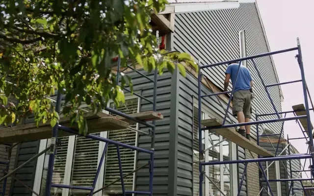 Un ouvrier du bâtiment sur un échafaudage installe un revêtement gris sur une maison.