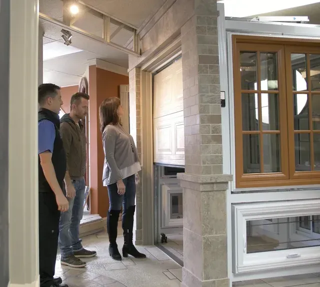 Trois personnes regardent une porte de garage exposée dans une salle d'exposition. Une femme blonde en bottes, des hommes en tenue décontractée.