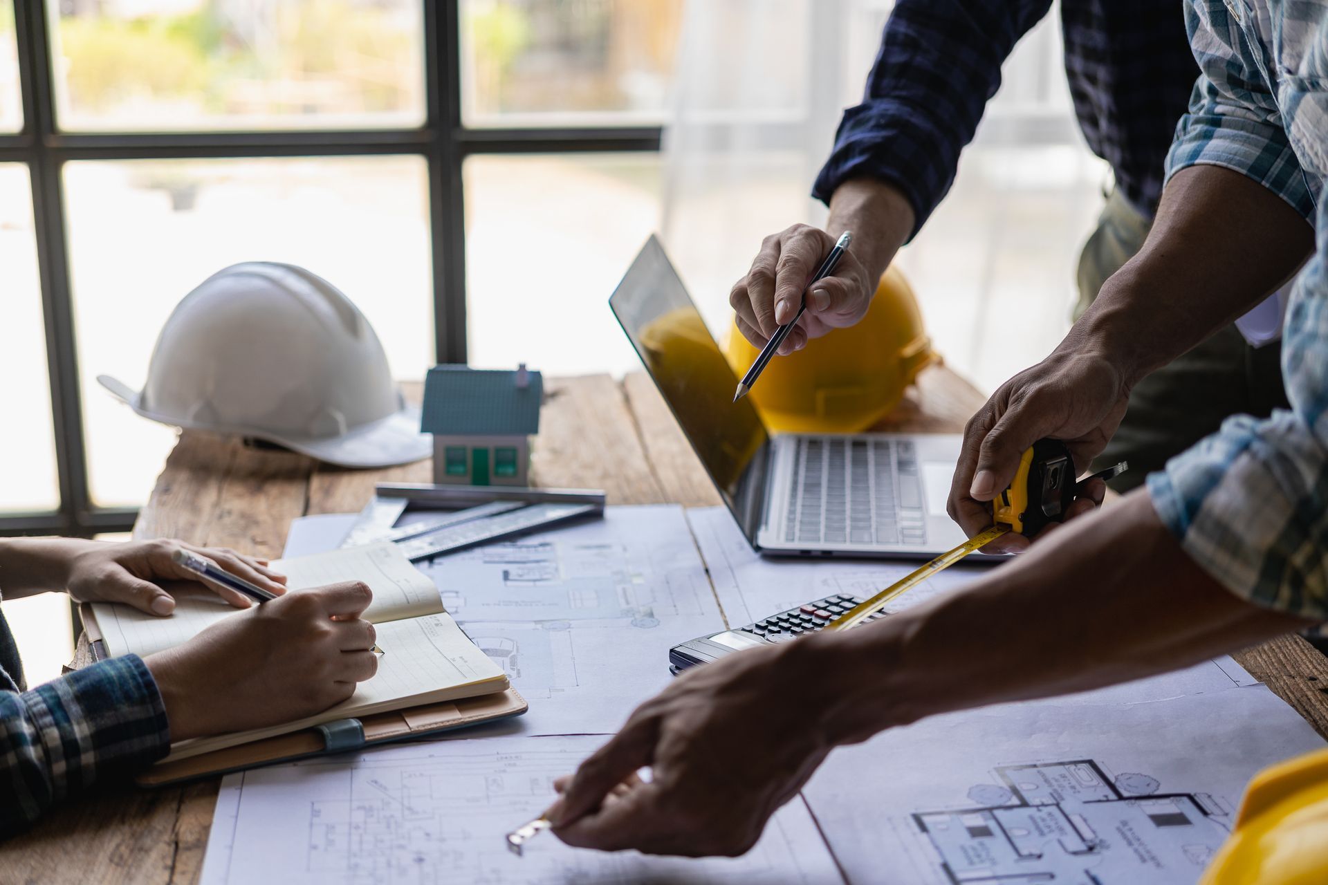 Des ouvriers du bâtiment examinent des plans à un bureau avec un ordinateur portable, des casques de sécurité et une maison modèle.