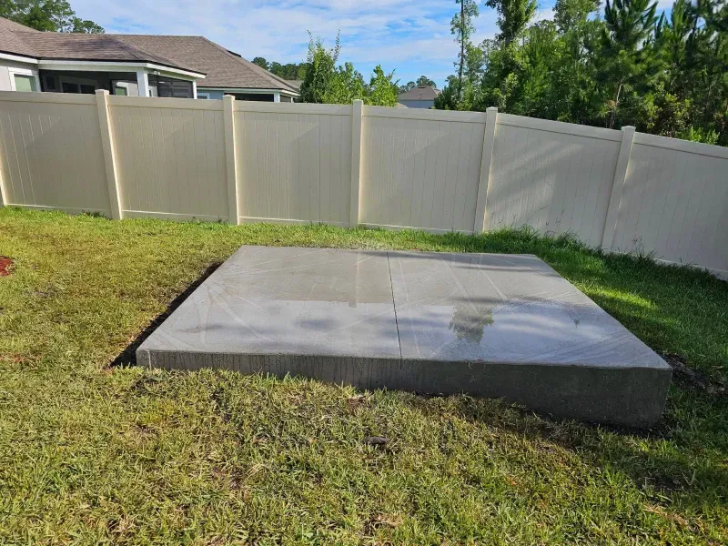 Concrete pad in a grassy backyard, surrounded by a white fence and a house.