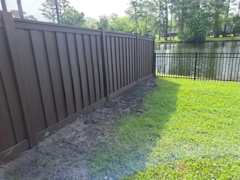 Brown privacy fence next to a black metal fence, overlooking a pond. Green grass in foreground.
