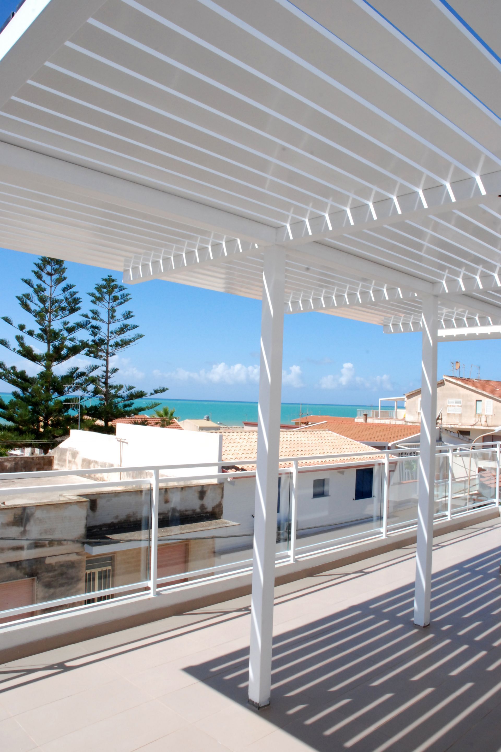 Terrazza con pergola bianca affacciata sui tetti del lungomare e sull'acqua azzurra sotto un cielo limpido