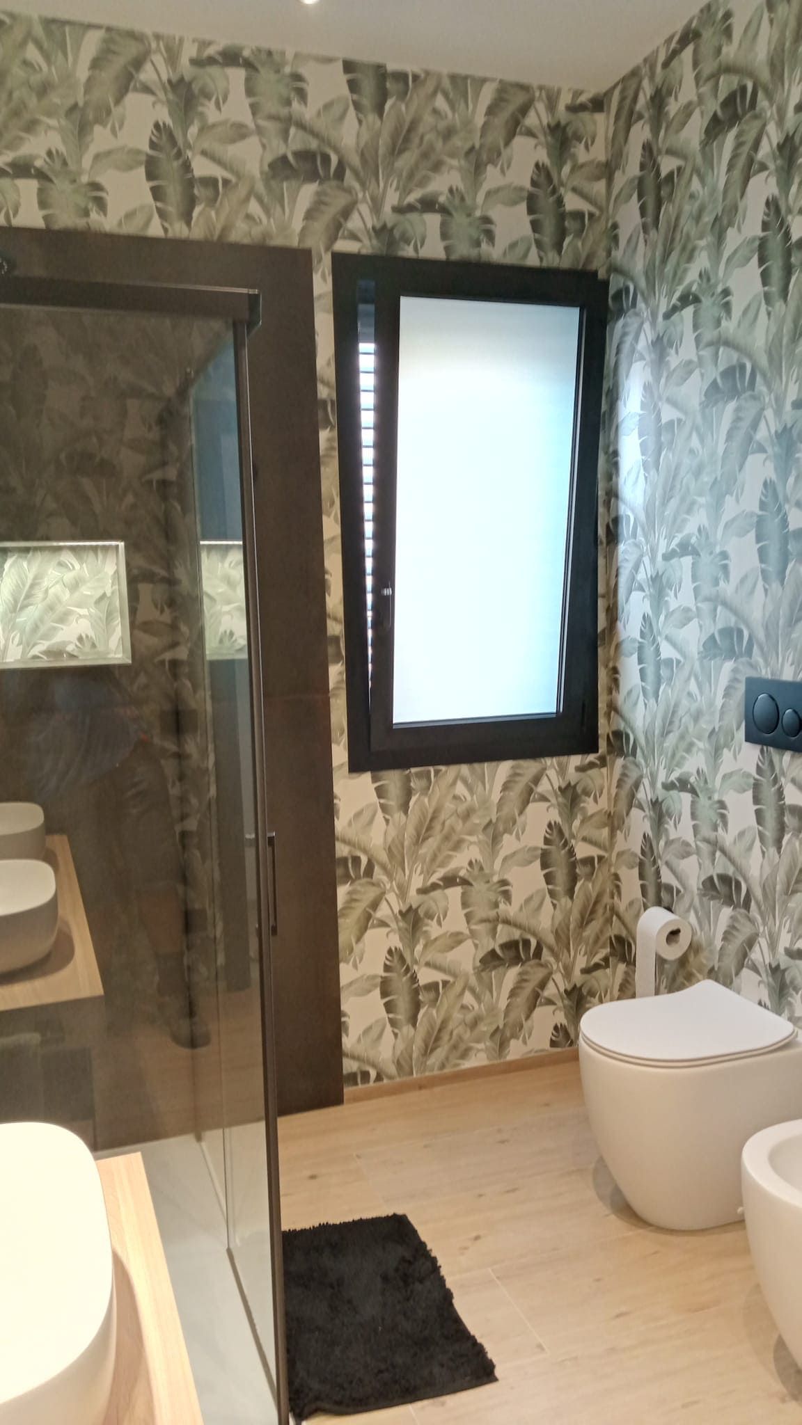 Bagno moderno con carta da parati a motivi geometrici, doccia in vetro, WC bianco e finestra satinata.
