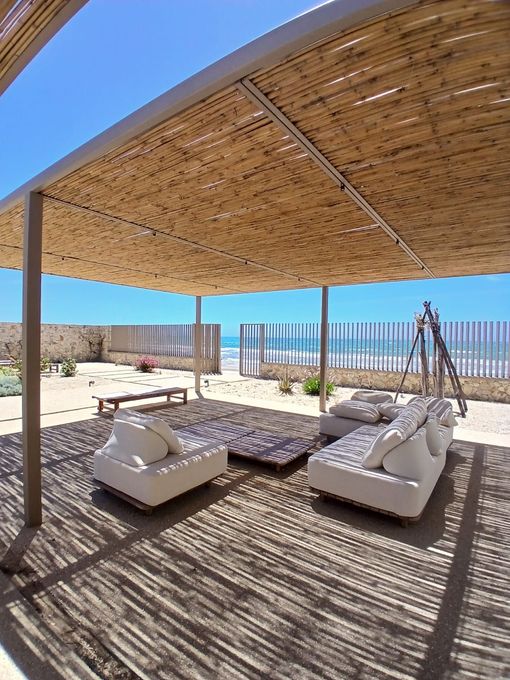 Patio sulla spiaggia ombreggiato con lettini bianchi, gazebo in vimini e vista sull'oceano.