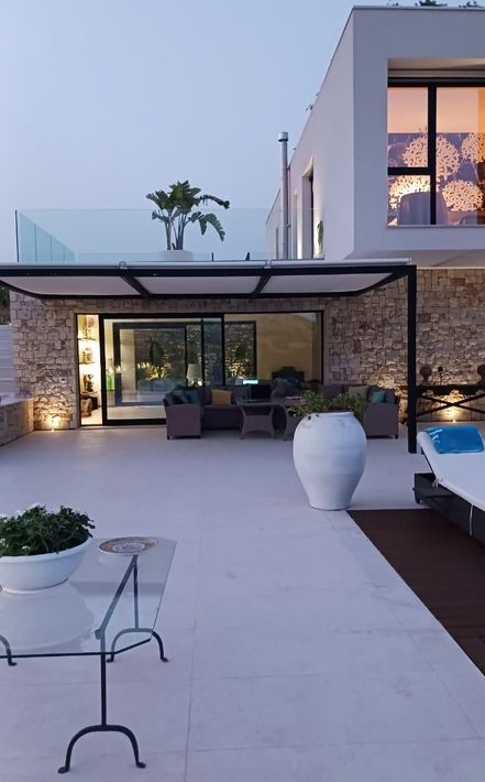 Cortile moderno con patio in pietra, posti a sedere all'aperto, grande vaso bianco, bordo piscina e casa illuminata al crepuscolo.