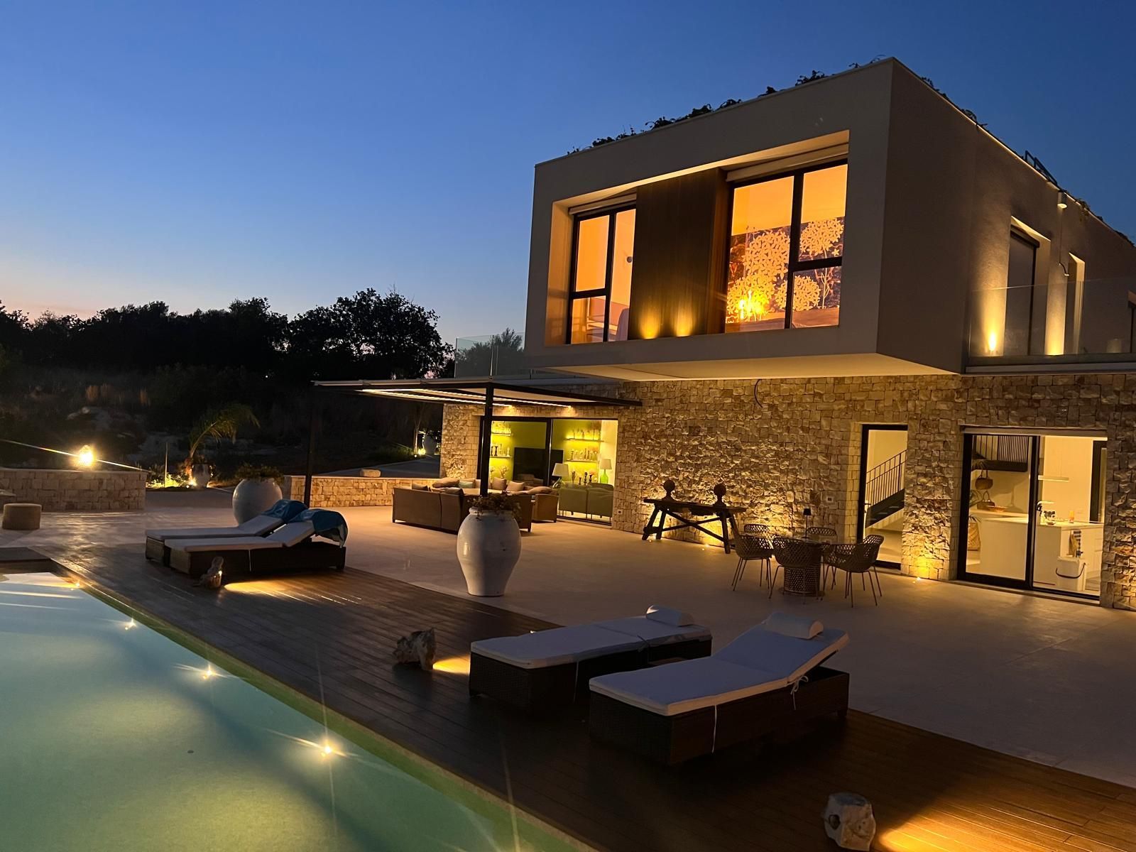 Lussuosa villa moderna al crepuscolo con finestre illuminate, lettini da esterno e una piscina che riflette il cielo serale