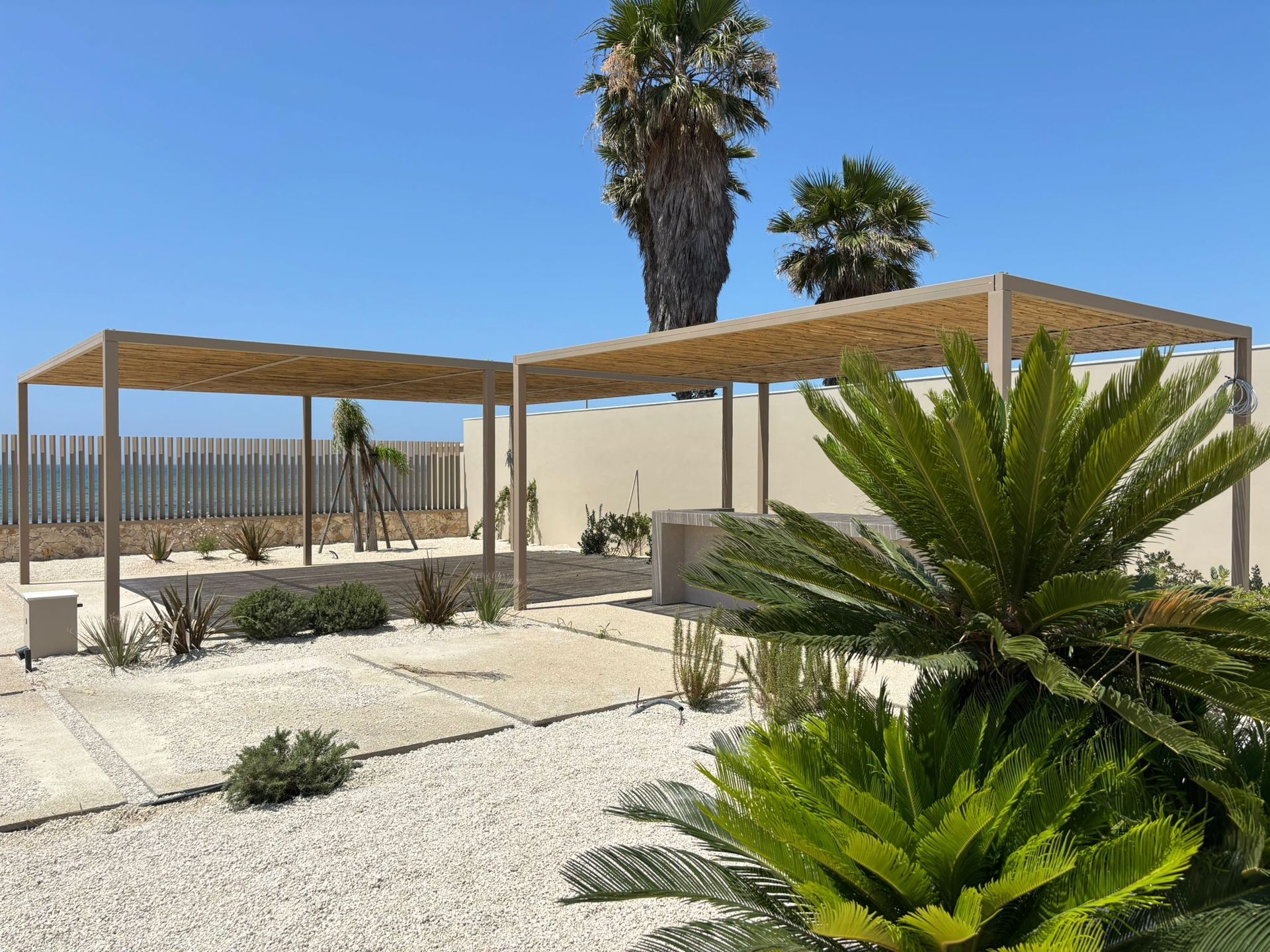 Patio nel deserto con pergole in legno, palme e ghiaia bianca sotto un cielo azzurro limpido.