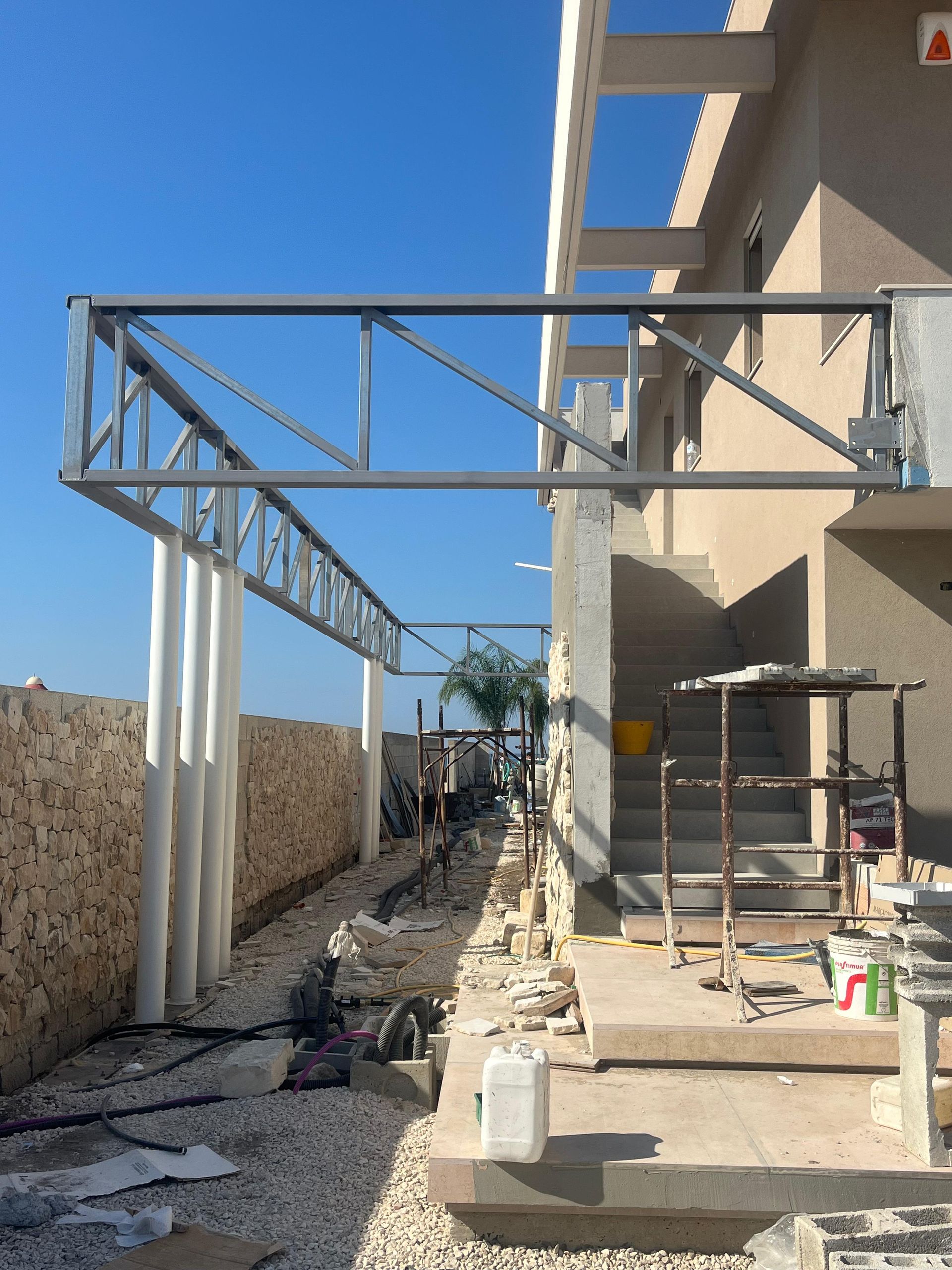 Cantiere edile all'aperto con struttura metallica per pergolato accanto a una casa beige e un muro in pietra sotto un cielo azzurro.