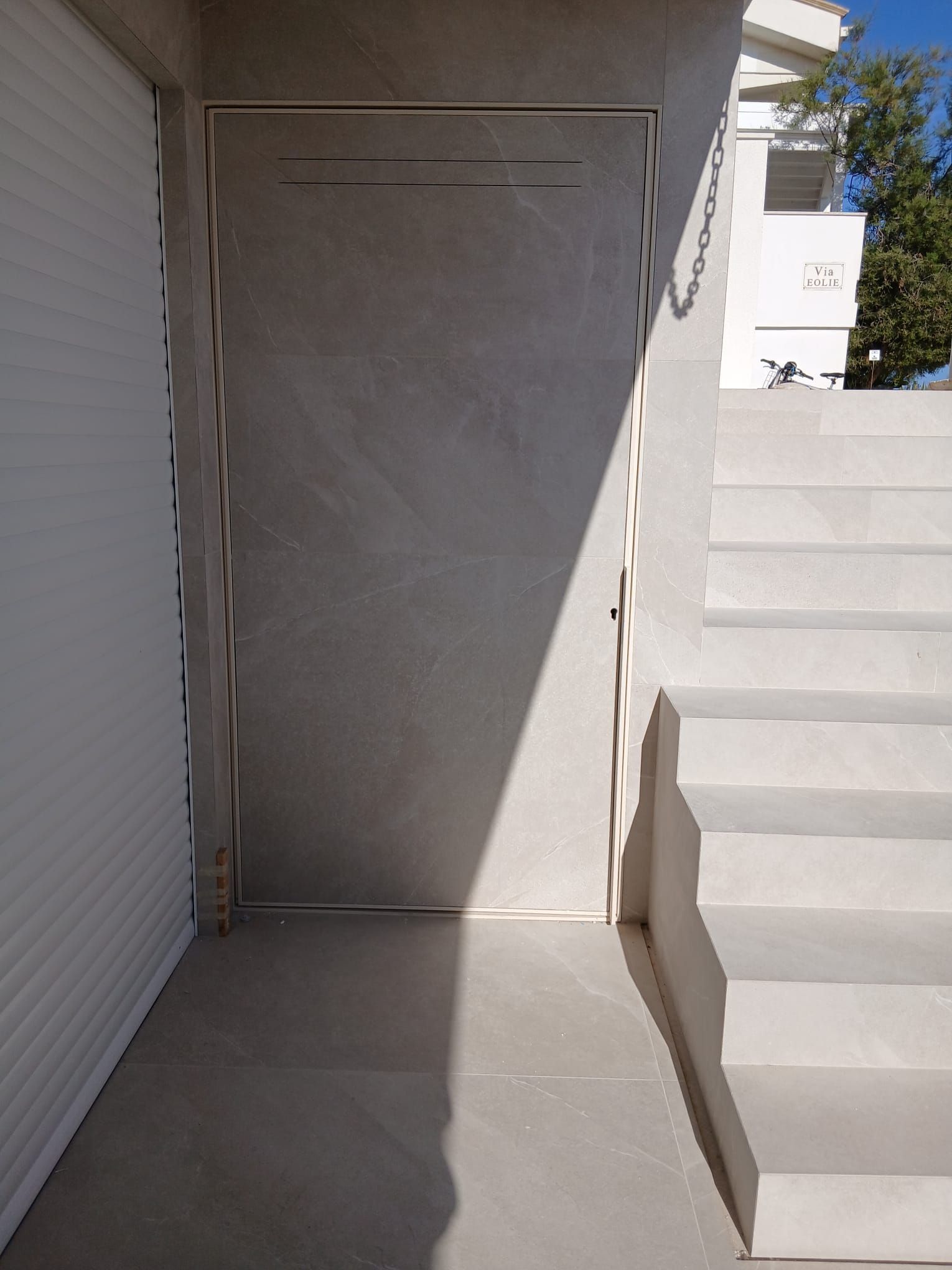 Stretto passaggio pedonale in cemento accanto a un muro in stucco e a una porta avvolgibile, con luce solare e scalini che conducono verso l'alto sulla destra.