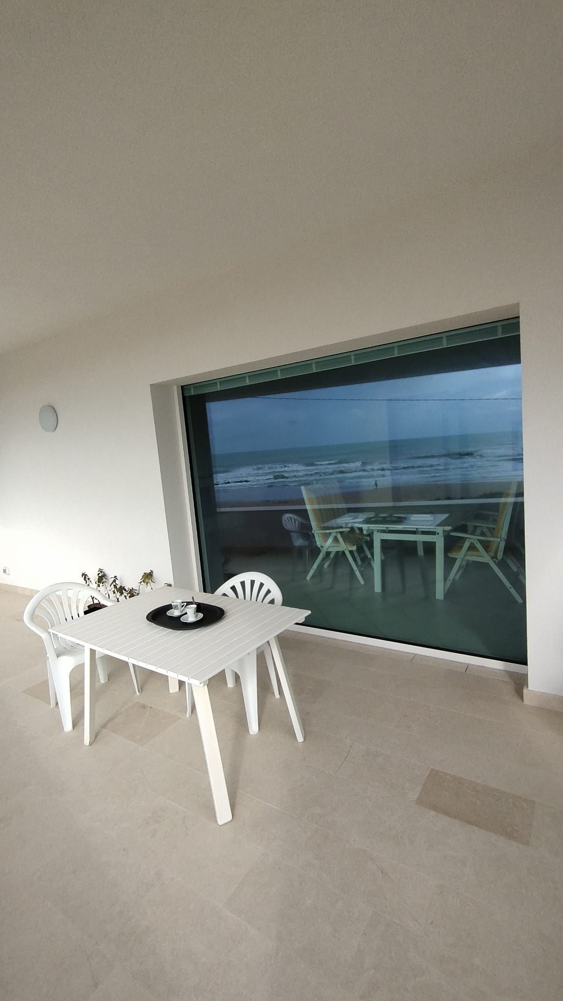 Patio coperto con tavolo e sedie bianchi, con vista sul mare attraverso un'ampia porta scorrevole in vetro.
