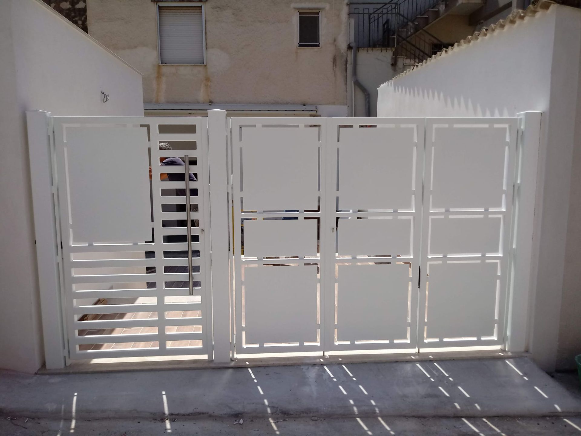 Cancello e recinzione in metallo bianco all'ingresso di un cortile soleggiato, con un edificio alle spalle.