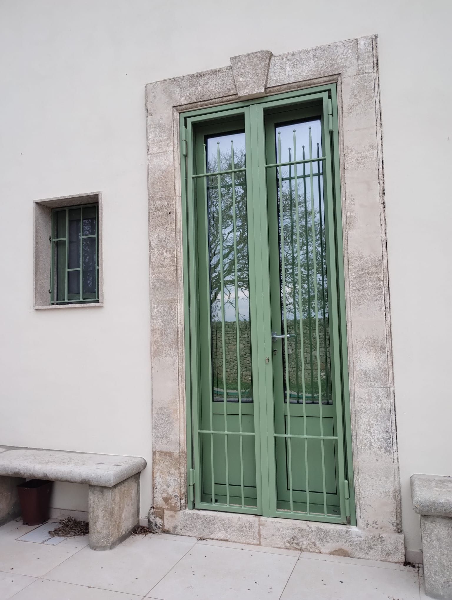 Porta metallica verde con piccola finestra a sbarre accanto a una finestra identica su una parete esterna intonacata.