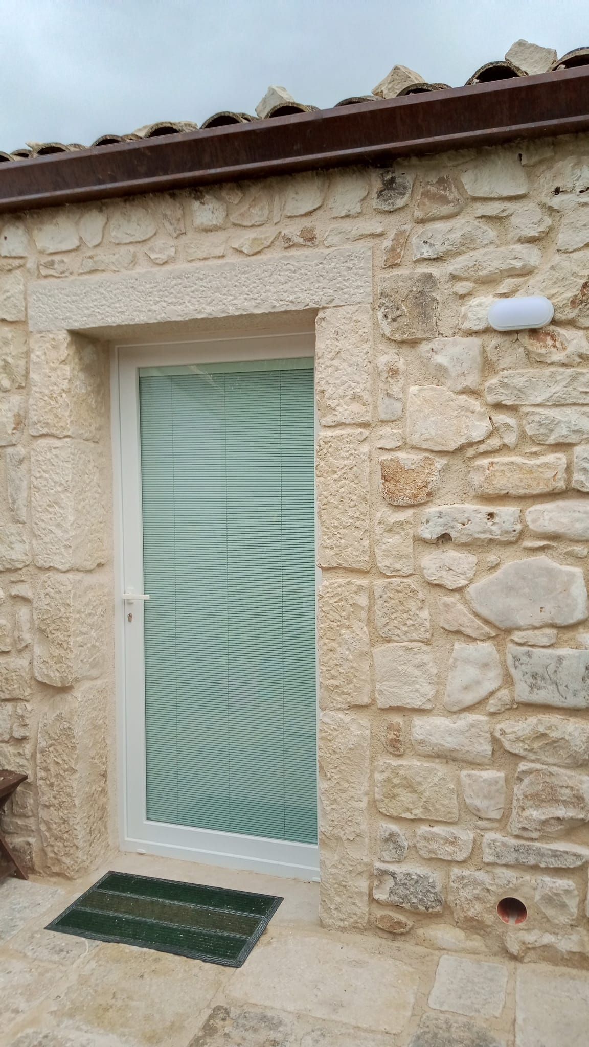 Esterno di una casa in pietra con porta in vetro satinato, zerbino verde e tetto di tegole.