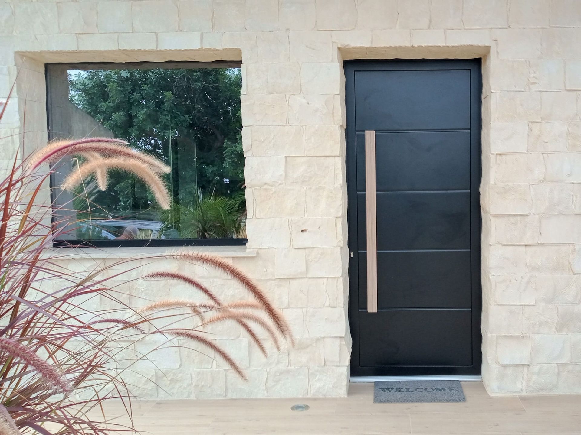 Porta d'ingresso nera moderna con maniglia lunga accanto a una finestra su una facciata di casa in pietra beige