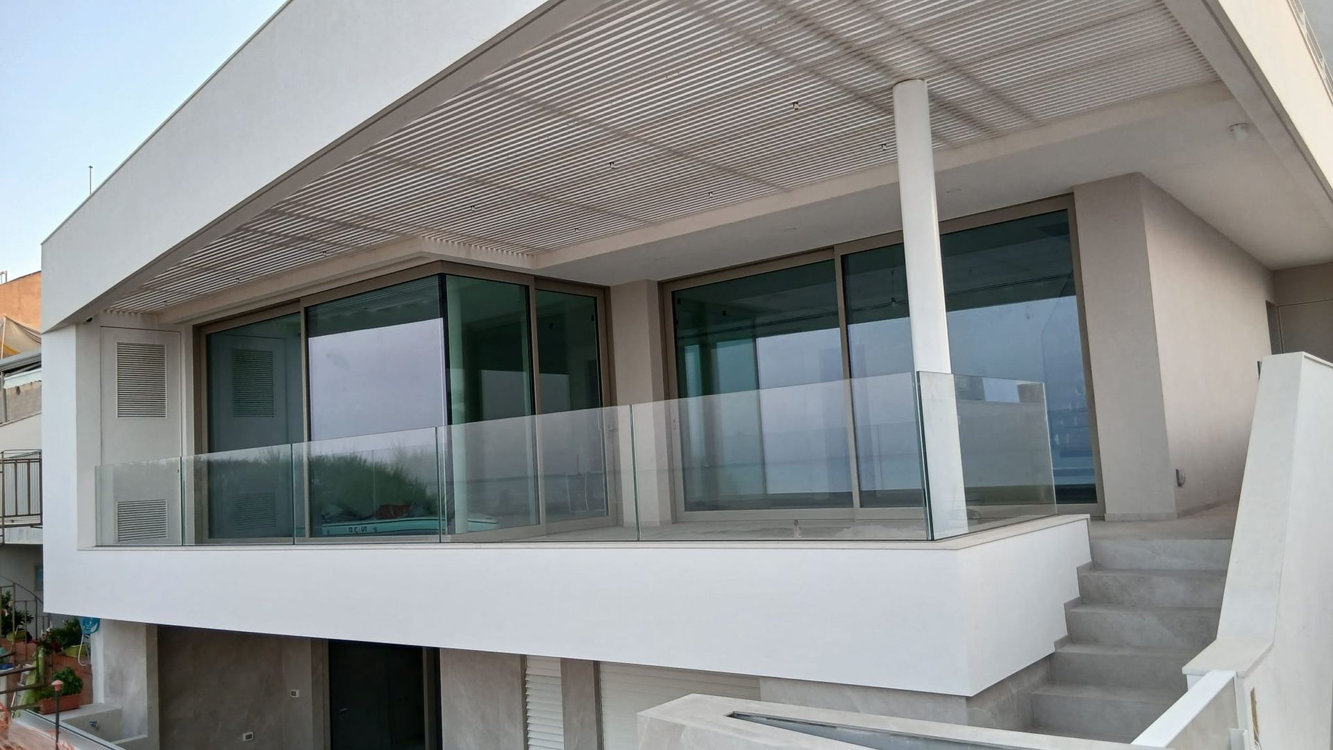 Balcone di una casa moderna con ringhiera in vetro e scale bianche sotto un pergolato ombreggiato.