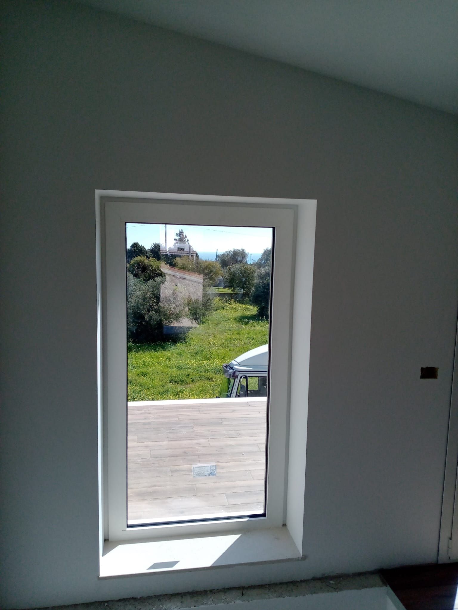 Interno di una stanza bianca con una porta aperta che incornicia una soleggiata vista esterna su prato, terrazza e alberi.