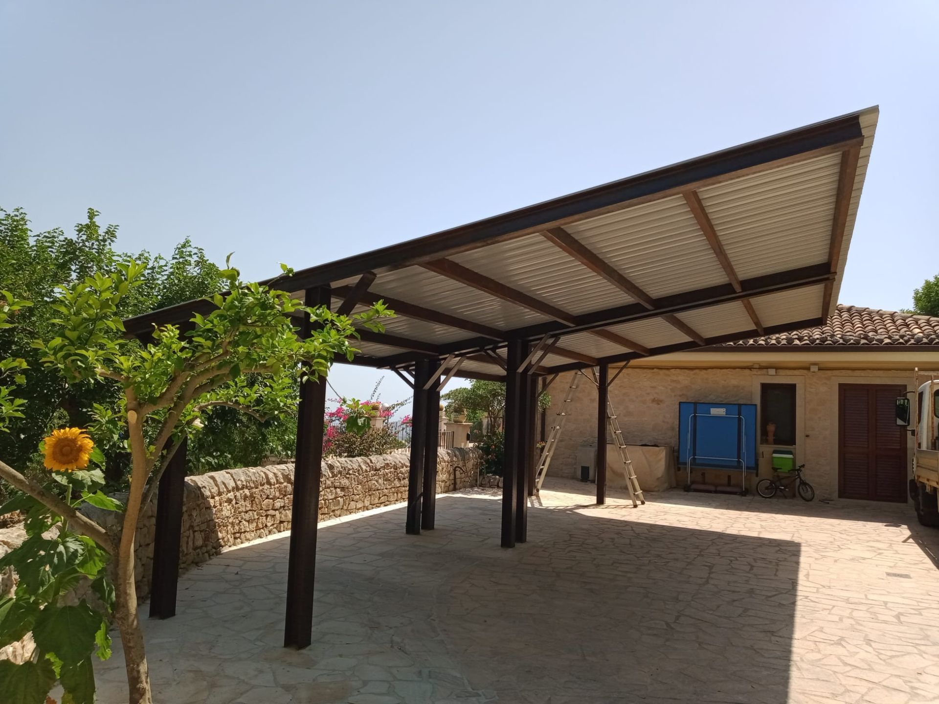 Patio coperto con tetto in metallo, muro in pietra e porta blu in un cortile soleggiato