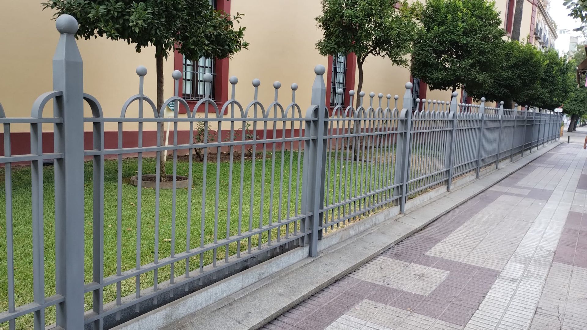 Una recinzione metallica grigia costeggia un marciapiede erboso accanto ad alberi e un edificio.