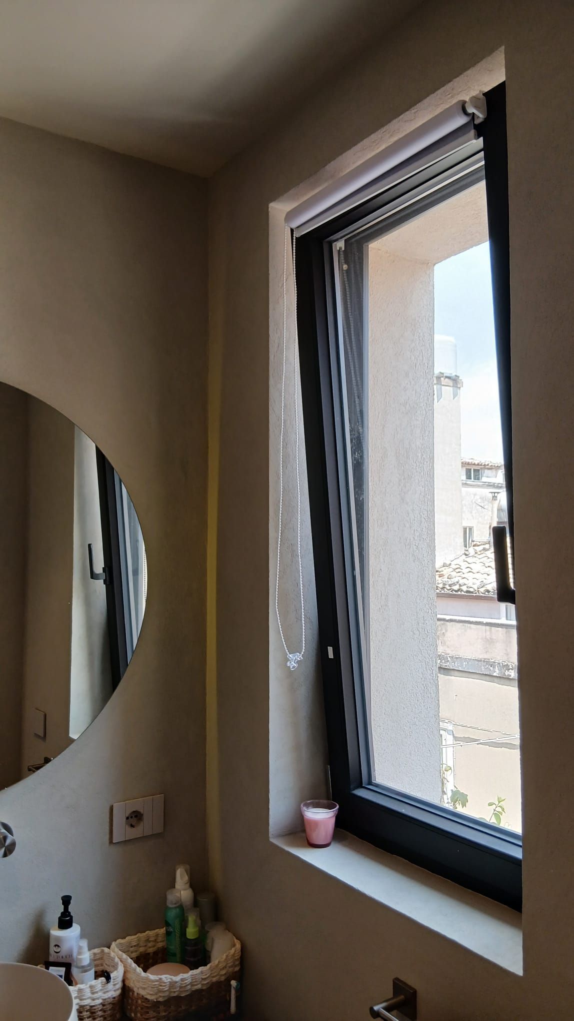 Angolo del bagno con specchio ovale, pareti beige e finestra con cornice nera sopra il ripiano del lavabo.