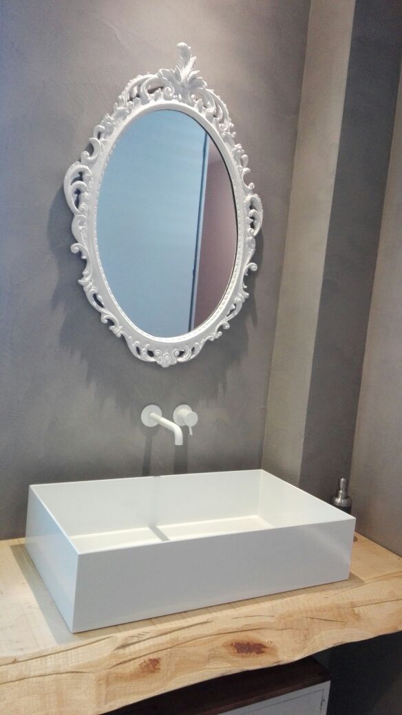 Mobile bagno bianco con specchio ovale decorato e rubinetto su piano in legno, contro una parete grigia.
