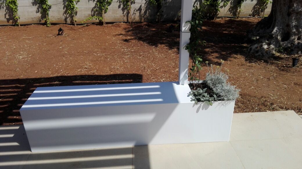 Panchina in cemento bianco in un cortile soleggiato e curato, con ombre a strisce e una piccola fioriera a un'estremità.