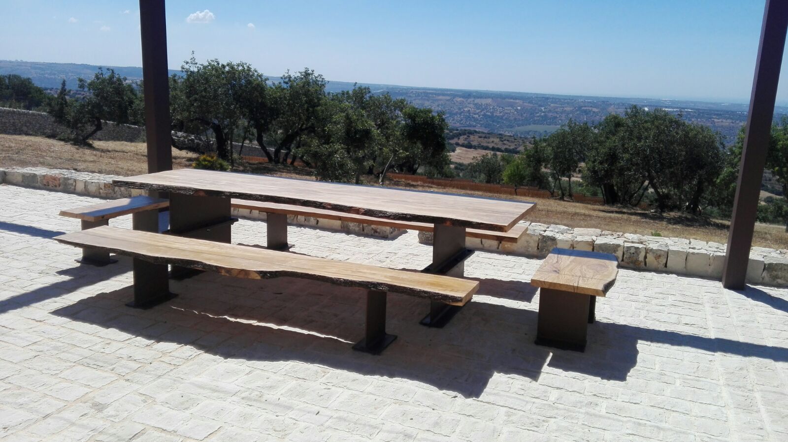 Tavolo e panche da picnic in legno su un patio soleggiato con vista su un paesaggio in lontananza.