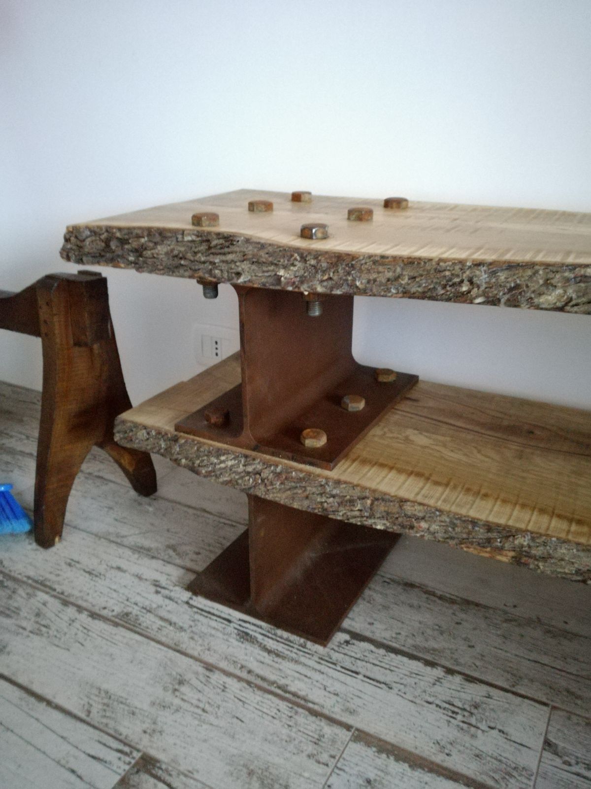 Panca in legno con ripiani a bordo naturale e piccoli pioli su un pavimento rustico dipinto di bianco.